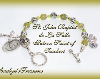 May include: A green and silver bracelet with a silver cross charm, a silver medal with a man's face, and a silver charm with a heart and the letter J. The bracelet also has a silver toggle clasp. The text on the bracelet reads "St. John Baptist de La Salle Patron Saint of Teachers".