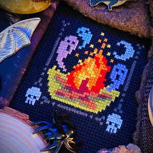 May include: A black cross-stitch pattern with a campfire, ghosts, and skulls. The campfire is made of orange, yellow, and red yarn. The ghosts are white and blue. The skulls are white.