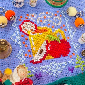 May include: A cross-stitch pattern on a purple fabric with a red, yellow, and white design of a person holding a mug. The pattern is surrounded by small white ceramic mugs, a small gold bell, and a small white and gold ornament.