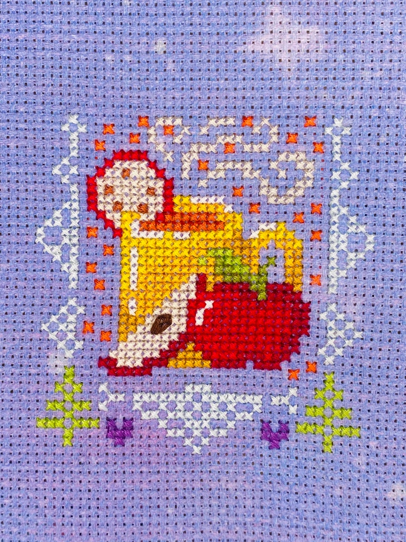 May include: Cross-stitch pattern of a mug of hot beverage with steam rising from it, a red apple, and orange and yellow accents on a purple background.