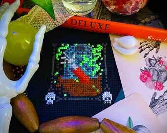 May include: A dark, atmospheric still life. A black cross-stitch design with a spooky motif is central. It's surrounded by a skeleton hand, wooden beads, books, and bottles. The color scheme is predominantly dark, with accents of green and orange.