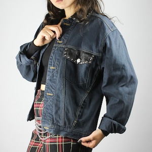 May include: A person wearing a dark blue denim jacket with silver studs, red and black checked trousers, and black boots. They are also wearing a black fedora hat and clear framed glasses.