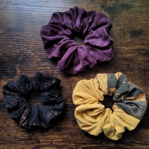 Könnte beinhalten: Nahaufnahme von drei Haargummis. Eines ist aus tiefviolettem Samt, ein anderes ist schwarz mit einem gesprenkelten Muster, und das dritte ist gelb mit einem grauen und gemusterten Design. Sie liegen auf einer dunklen Holzoberfläche.