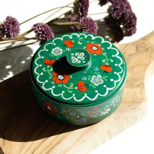 May include: A round green wooden box with a lid featuring a hand-painted floral pattern in red, white, and blue. The lid has a central knob and a scalloped white border design. The sides of the box are also decorated with small painted flowers.