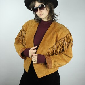 May include: A brown suede jacket with fringe detailing on the sleeves and collar. The jacket has a button closure and a pointed collar. The model is wearing a black hat and sunglasses.