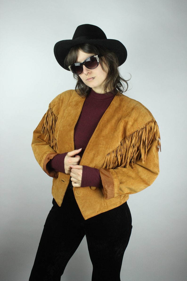 May include: A brown suede jacket with fringe detailing on the sleeves and collar. The jacket has a button closure and a pointed collar. The model is wearing a black hat and sunglasses.