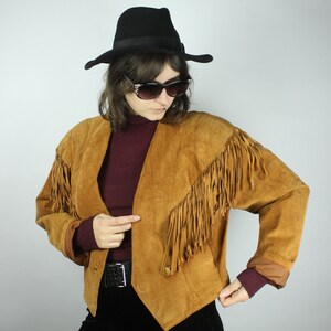 May include: A brown suede jacket with fringe detailing. The jacket has a pointed collar and a button closure. The model is wearing a burgundy turtleneck and black trousers.