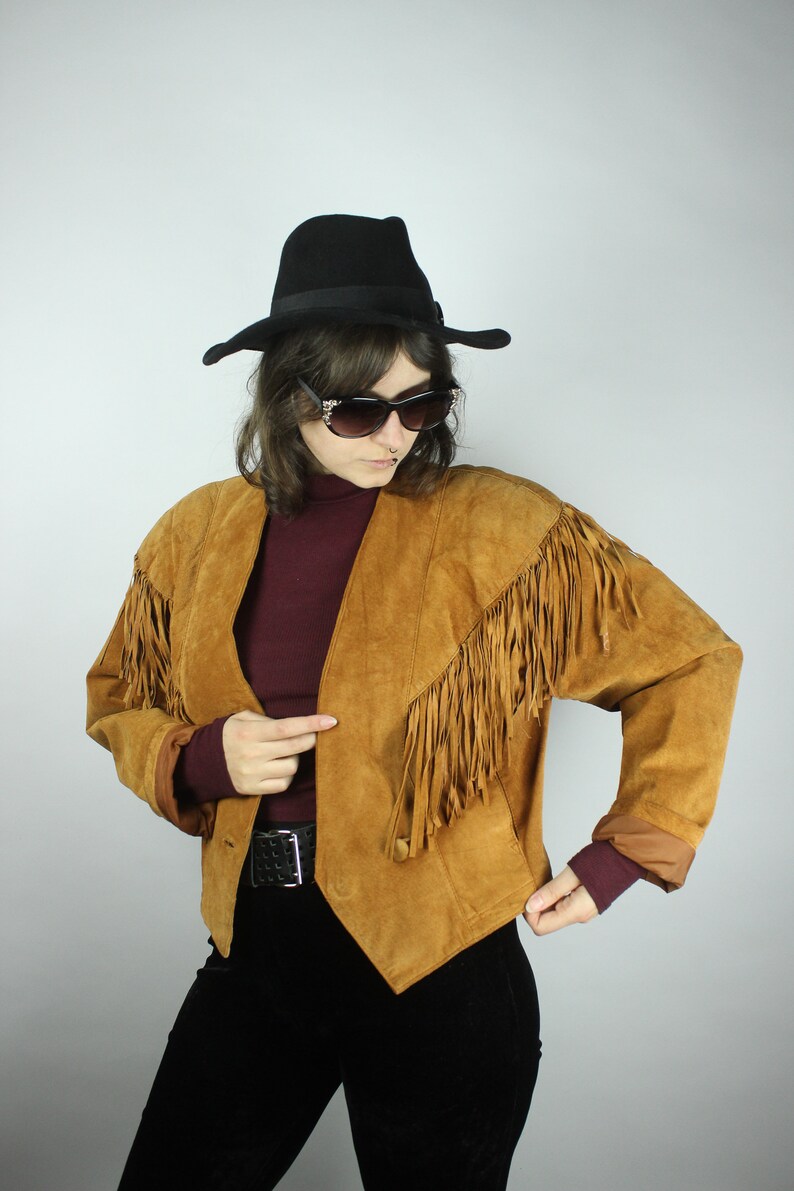 May include: A brown suede jacket with fringe detailing. The jacket has a pointed collar and a button closure. The model is wearing a burgundy turtleneck and black trousers.