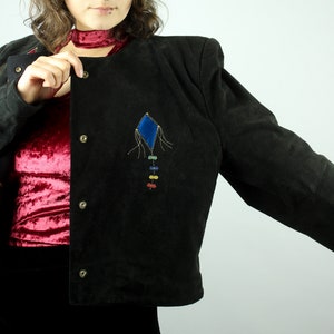 May include: A black suede jacket with a blue and rainbow kite embroidered on the front. The jacket has a button closure and a collar.