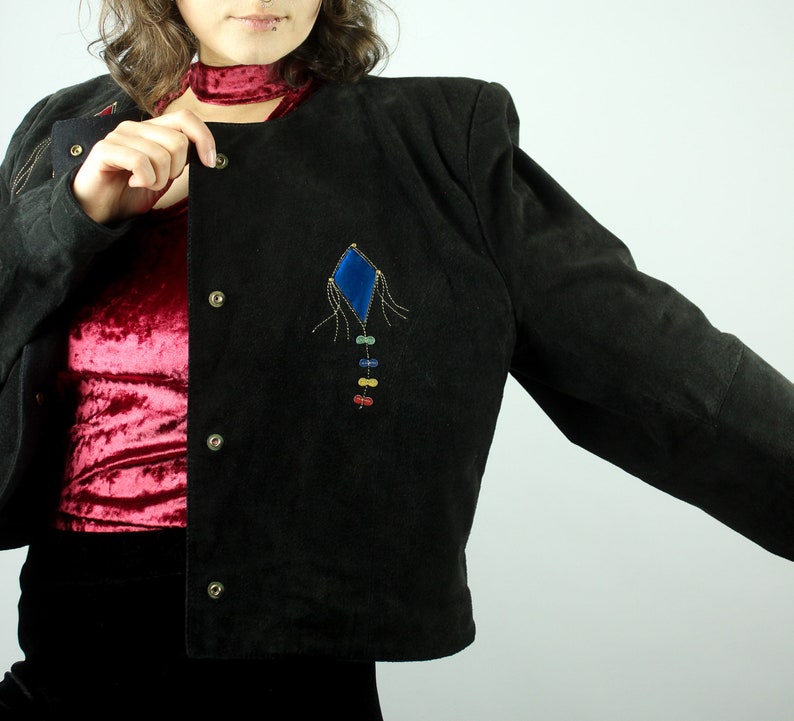 May include: A black suede jacket with a blue and rainbow kite embroidered on the front. The jacket has a button closure and a collar.