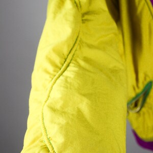 May include: Close-up of a yellow and purple jacket sleeve. The sleeve is made of a smooth, shiny fabric and has a green seam running down the side. The sleeve is cuffed with a purple band.