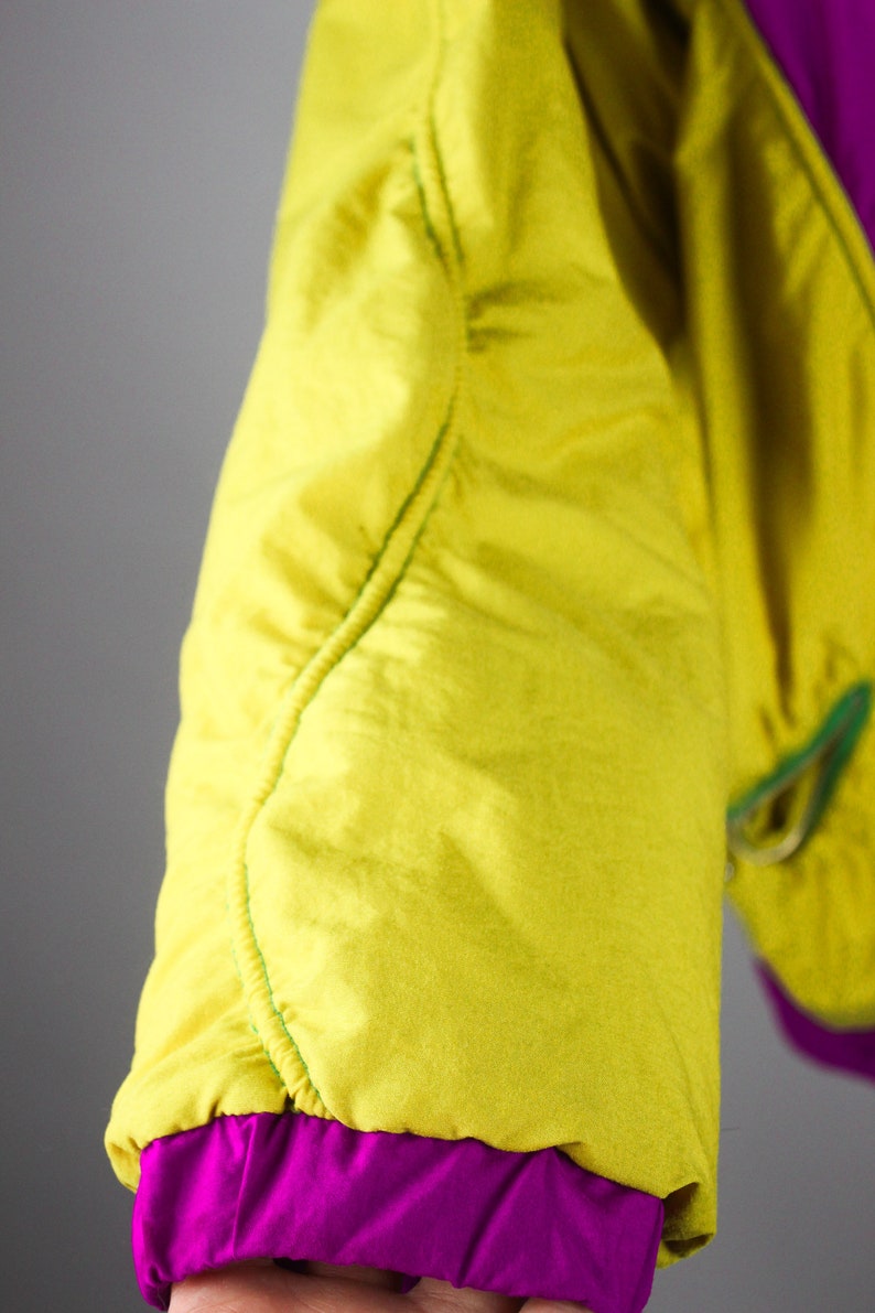 May include: Close-up of a yellow and purple jacket sleeve. The sleeve is made of a smooth, shiny fabric and has a green seam running down the side. The sleeve is cuffed with a purple band.