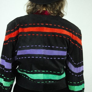 May include: A black suede jacket with colorful striped detailing. The jacket has red, purple, and green stripes with black stitching. The jacket is worn by a person wearing a black hat.