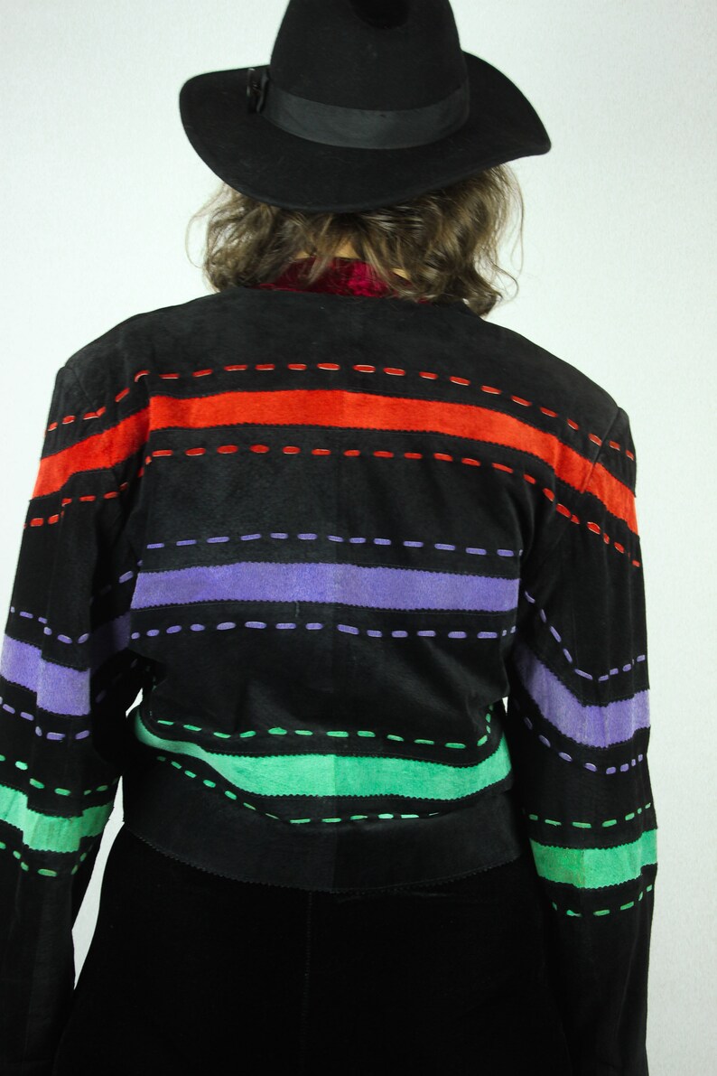 May include: A black suede jacket with colorful striped detailing. The jacket has red, purple, and green stripes with black stitching. The jacket is worn by a person wearing a black hat.