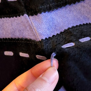 May include: A close-up of a purple leather cord being tied through a series of holes in a black leather surface. The leather is stitched with a decorative stitch.