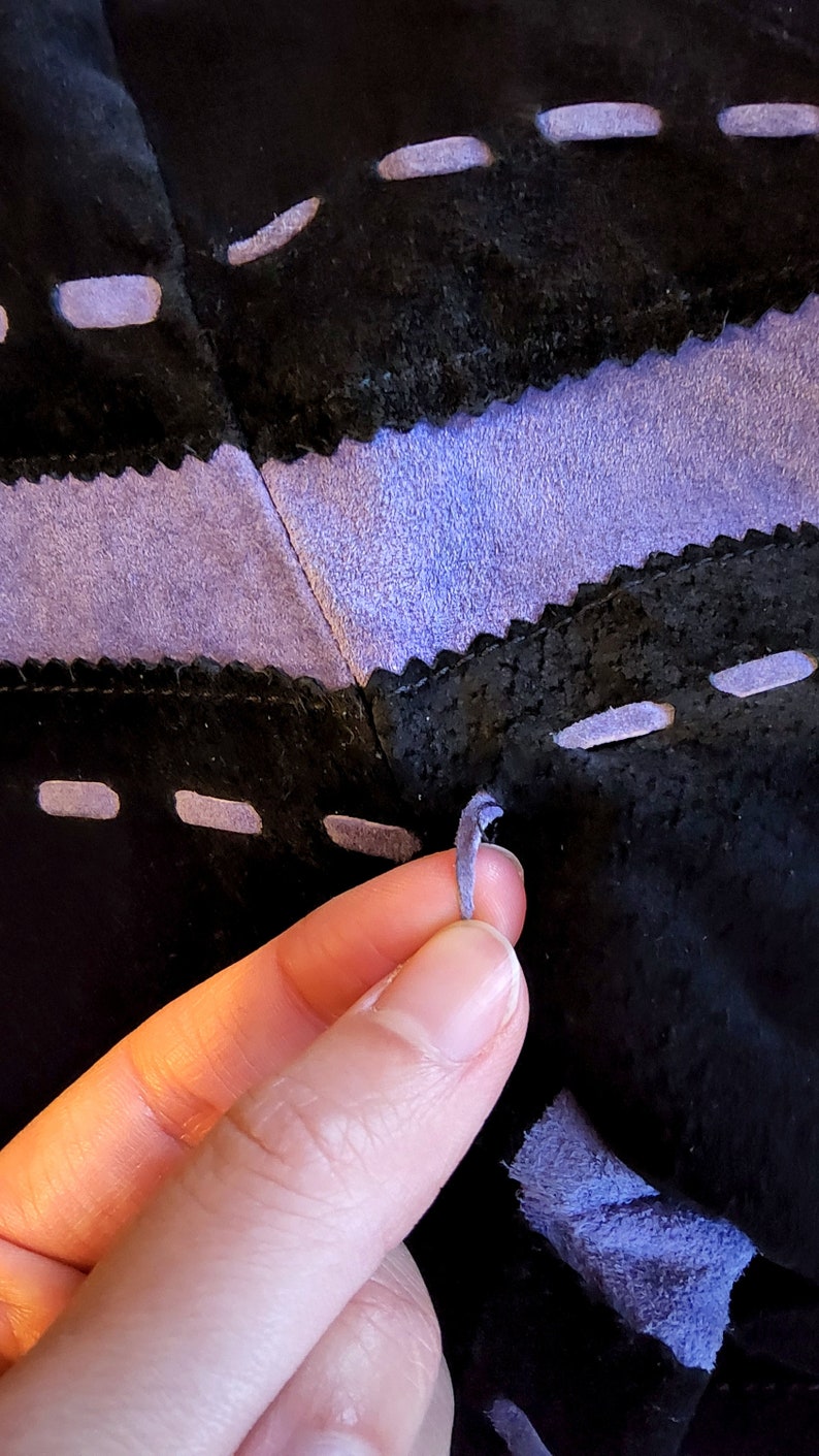 May include: A close-up of a purple leather cord being tied through a series of holes in a black leather surface. The leather is stitched with a decorative stitch.