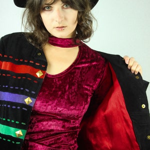 May include: A woman wearing a black corduroy jacket with a red lining, a burgundy velvet top, and a black hat. The jacket has colorful stripes and gold accents.