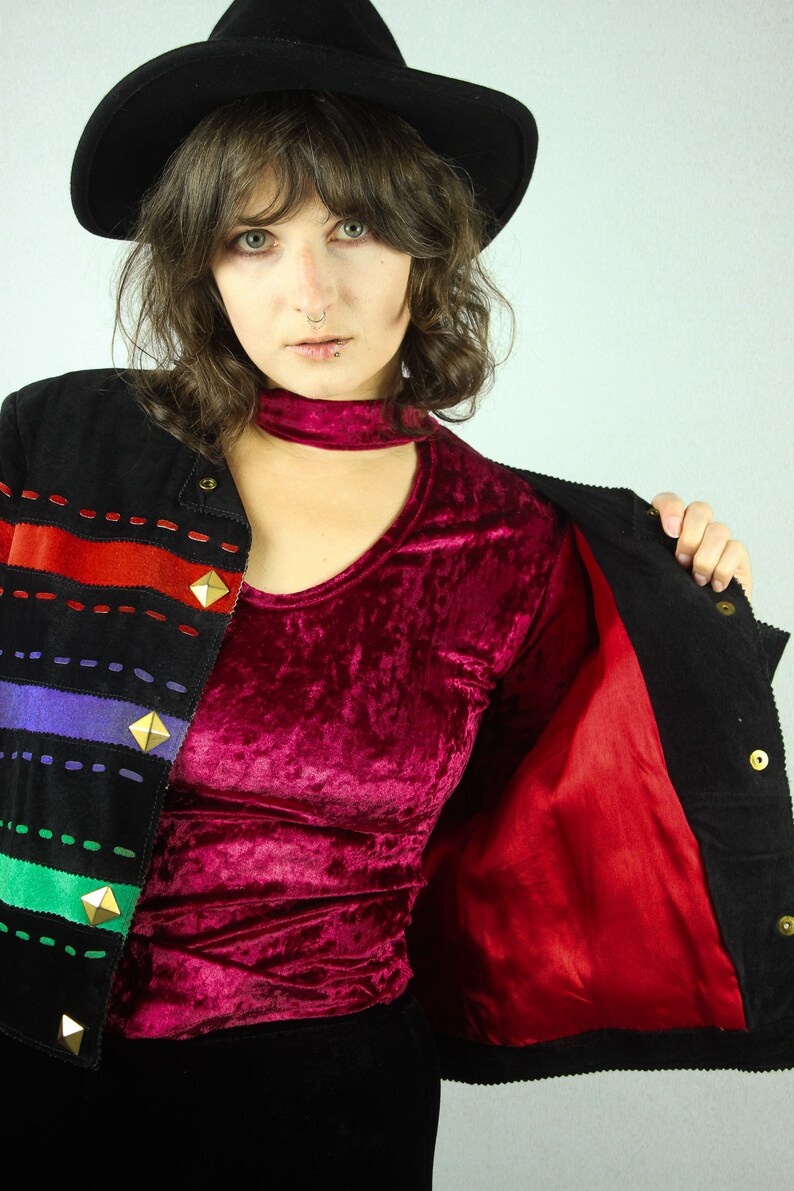 May include: A woman wearing a black corduroy jacket with a red lining, a burgundy velvet top, and a black hat. The jacket has colorful stripes and gold accents.