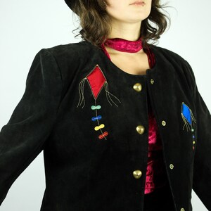 May include: A black suede jacket with a red and blue diamond-shaped design embroidered on the front. The jacket has gold buttons and a red velvet collar.