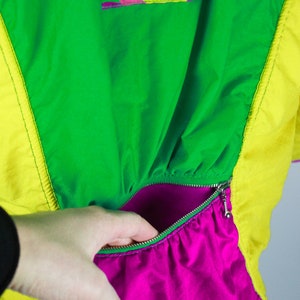 May include: A bright green, yellow and pink jacket with a zipped pocket. The pocket is lined with a bright pink fabric. The jacket has a graphic on the front that says "Camp Out" with a peace sign.