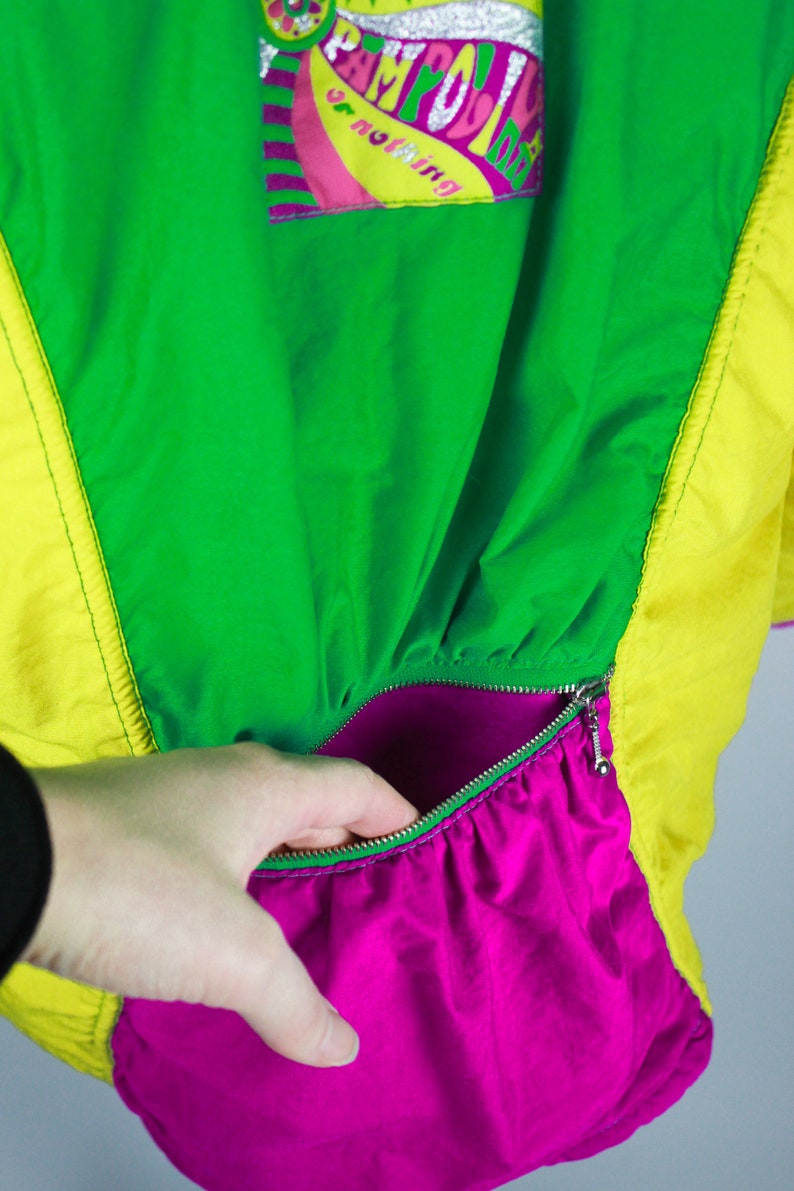 May include: A bright green, yellow and pink jacket with a zipped pocket. The pocket is lined with a bright pink fabric. The jacket has a graphic on the front that says "Camp Out" with a peace sign.