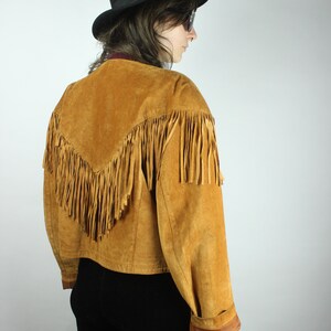 May include: A brown suede jacket with fringe detailing. The jacket has a cropped fit and long sleeves. The model is wearing a black hat and sunglasses.