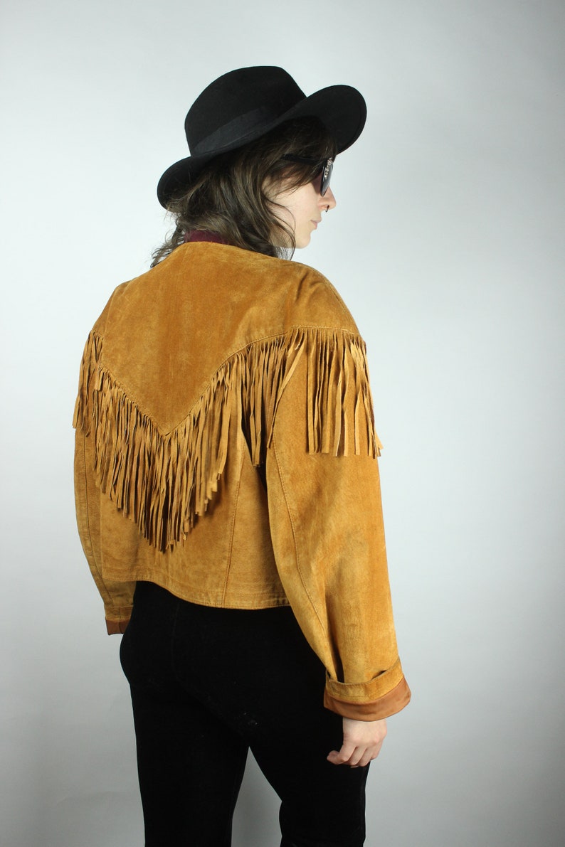 May include: A brown suede jacket with fringe detailing. The jacket has a cropped fit and long sleeves. The model is wearing a black hat and sunglasses.