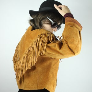 May include: A brown suede jacket with fringe detailing. The jacket has a collar and long sleeves. The model is wearing a black hat and sunglasses.