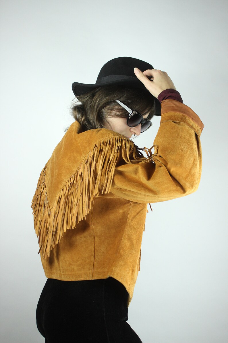 May include: A brown suede jacket with fringe detailing. The jacket has a collar and long sleeves. The model is wearing a black hat and sunglasses.