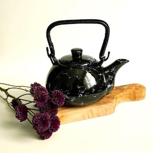 Peut inclure: Théière en céramique noire avec un motif marbré et une anse tressée. La théière est posée sur une planche en bois, avec des fleurs violettes au premier plan. Elle a un bec courbé et un couvercle.
