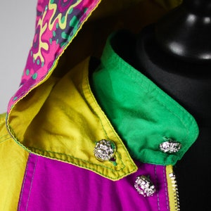 May include: A colourful jacket with a pink, yellow and green colour scheme. The jacket has a hood with a patterned design and a zip closure. The jacket has decorative buttons.