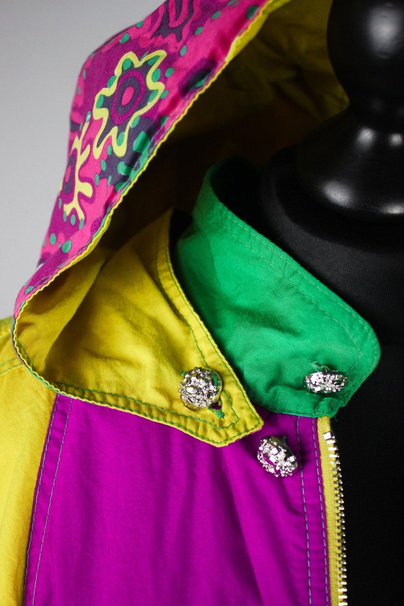 May include: A colourful jacket with a pink, yellow and green colour scheme. The jacket has a hood with a patterned design and a zip closure. The jacket has decorative buttons.