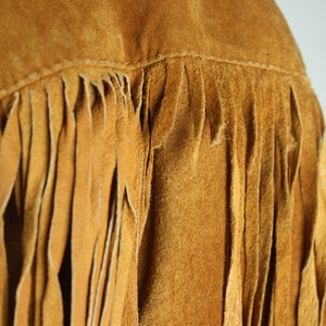 May include: Close-up of a brown suede jacket with fringe detailing. The fringe is made of the same material as the jacket and hangs down from the bottom edge.