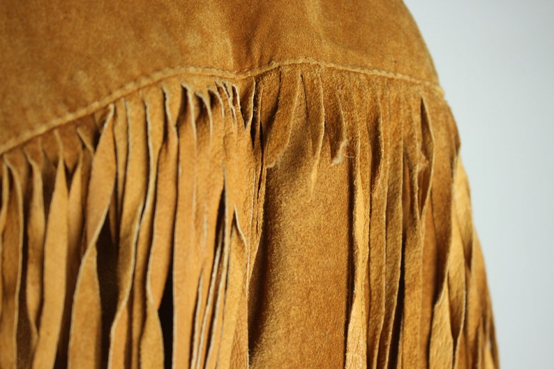 May include: Close-up of a brown suede jacket with fringe detailing. The fringe is made of the same material as the jacket and hangs down from the bottom edge.