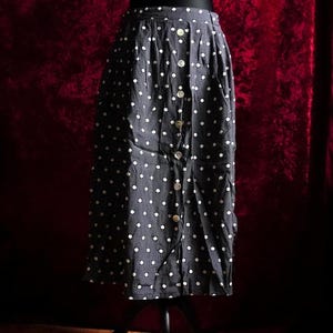 Könnte beinhalten: Marineblauer Rock mit weißen Polka Dots und Knopfleiste. Der Rock wird auf einer schwarzen Schaufensterpuppe präsentiert und hat eine hohe Taille und einen knielangen Saum. Die Knöpfe sind hellbeige.