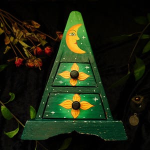 以下が含まれることがあります： 緑色の三角形の木製ジュエリーボックス。2つの引き出しがあり、上部には三日月と星、引き出しには花の模様が描かれています。引き出しのつまみは黒色です。背景は暗く、葉があります。