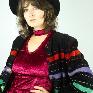 May include: A woman wearing a black hat, a red velvet top, and a black jacket with colorful stripes. The jacket has a zipper and a button closure.