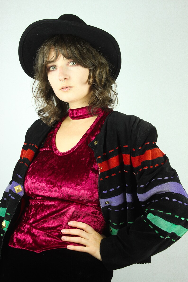 May include: A woman wearing a black hat, a red velvet top, and a black jacket with colorful stripes. The jacket has a zipper and a button closure.