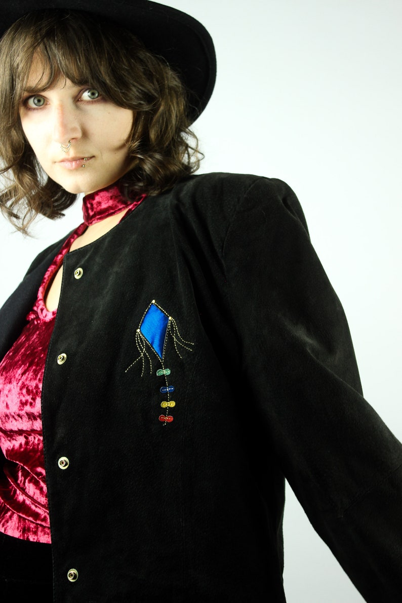 May include: A black suede jacket with a colourful kite design on the front. The jacket has a red velvet collar and a button closure.