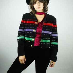May include: A woman wearing a black suede jacket with colorful stripes and gold buttons. The jacket has a red, green, purple, and blue striped pattern. She is also wearing black bell bottom pants and a black hat.