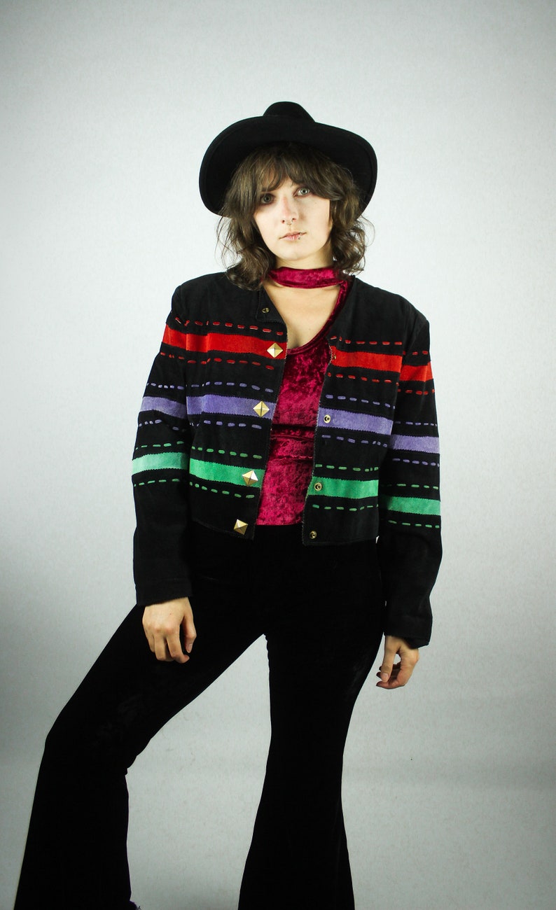 May include: A woman wearing a black suede jacket with colorful stripes and gold buttons. The jacket has a red, green, purple, and blue striped pattern. She is also wearing black bell bottom pants and a black hat.