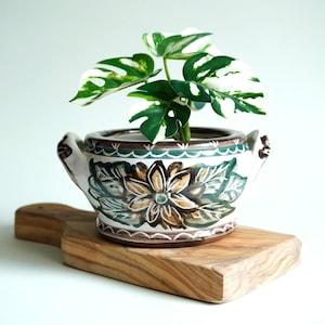 Könnte beinhalten: Ein Keramik-Blumentopf mit floralem Design, der eine Pflanze mit grün-weißen Blättern enthält. Der Topf steht auf einem Holzschneidebrett. Der Topf hat Griffe und ist braun, grün und weiß.