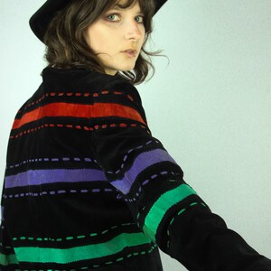May include: A black suede jacket with colorful striped stitching. The jacket has red, purple, and green stripes. The model is wearing a black hat.