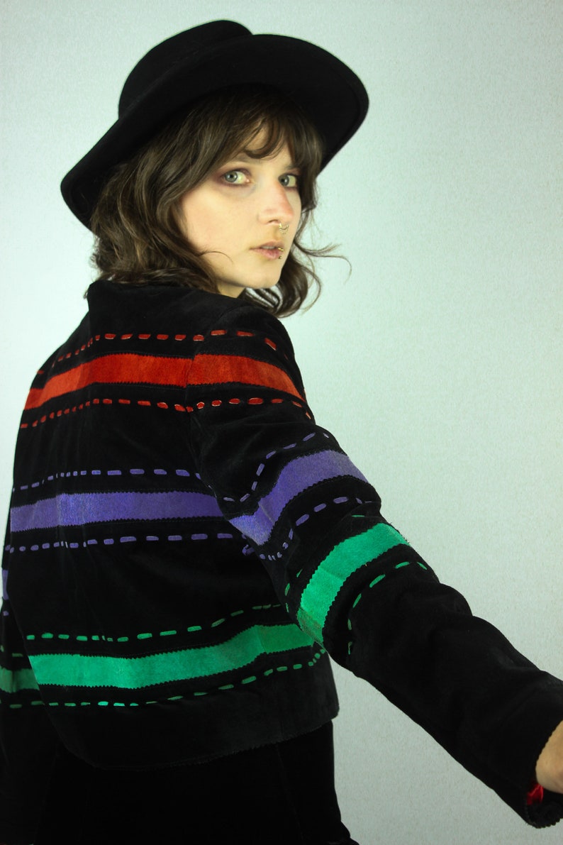May include: A black suede jacket with colorful striped stitching. The jacket has red, purple, and green stripes. The model is wearing a black hat.