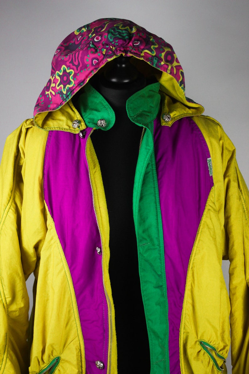 May include: A yellow, green and purple ski jacket with a colourful patterned hood. The jacket has a zip closure and multiple pockets.