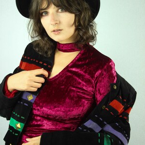 May include: A woman wearing a black velvet choker, a burgundy velvet top, and a black jacket with colorful stitching and gold accents. She is also wearing a black fedora.