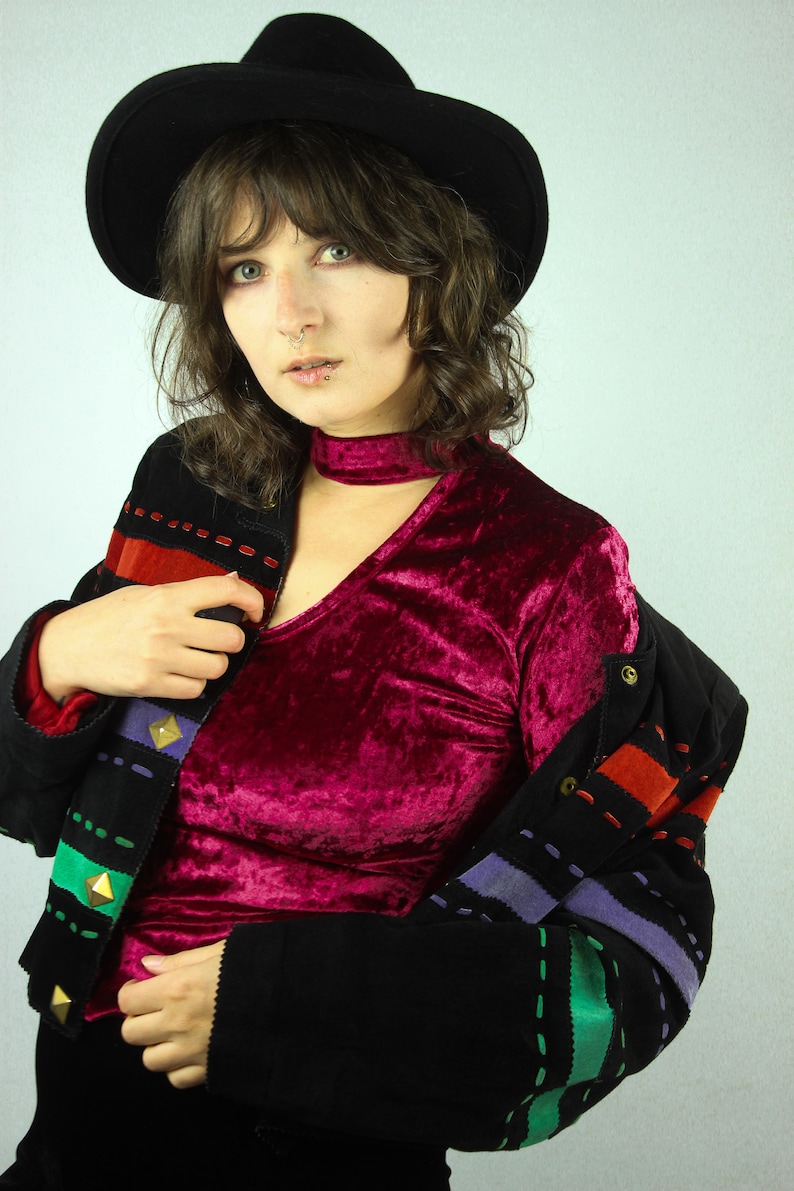 May include: A woman wearing a black velvet choker, a burgundy velvet top, and a black jacket with colorful stitching and gold accents. She is also wearing a black fedora.