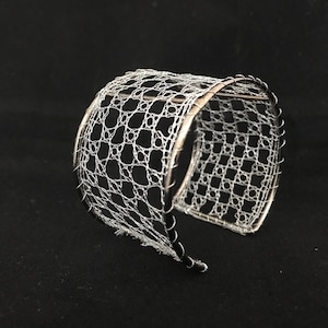 May include: A silver metal bracelet with a delicate lace design. The bracelet is open and has a slightly curved shape.