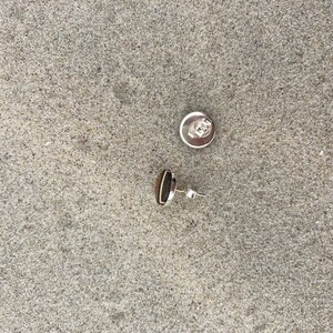 May include: A pair of silver stud earrings with a dark brown oval-shaped stone in the center.