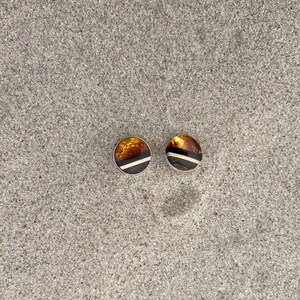 May include: A pair of amber and black striped earrings with silver settings. The earrings are on a sandy surface.